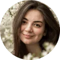 Smiling young woman with long brown hair outdoors with blurred white flowers in the background.