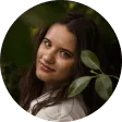 Woman with long brown hair and brown eyes looking at the camera, surrounded by green leaves.