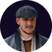 Man wearing a flat cap, dark jacket, scarf, and a brown shirt, smiling slightly against a dark background.