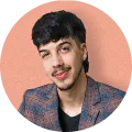Portrait of a man with dark hair and a mustache, wearing a checked blazer and black shirt, smiling slightly against a peach background.