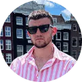 Man wearing black sunglasses and a pink striped shirt standing outdoors with brick buildings in the background.